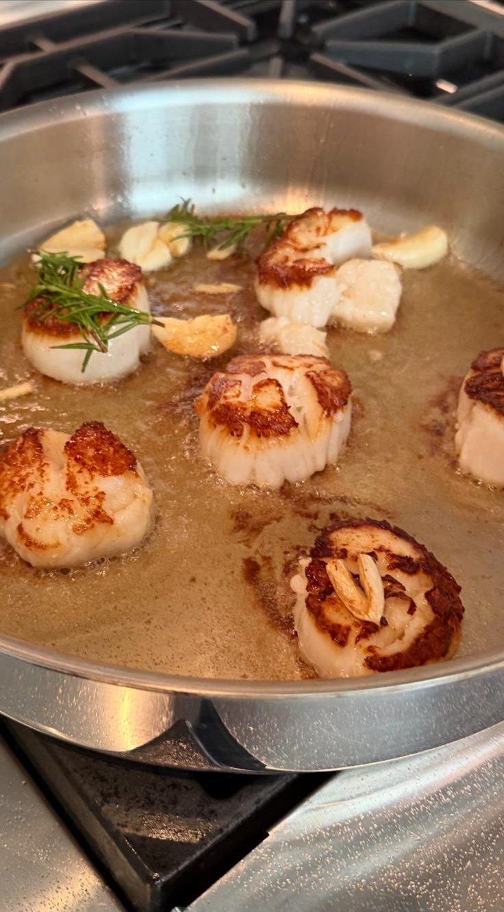 Scallops searing in a stainless pan with garlic and rosemary, golden crust on each side.